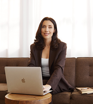 Annika Marshall sitting on a couch in a brown suit, smiling behind a laptop