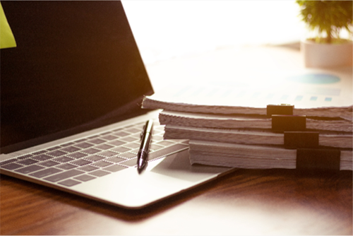 laptop with a stack of paperwork in front of it