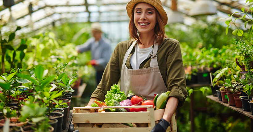 young woman standing in a plant nursery holding a crate of vegetables