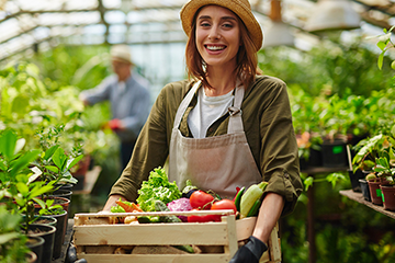 young woman standing in a plant nursery holding a box of vegetables